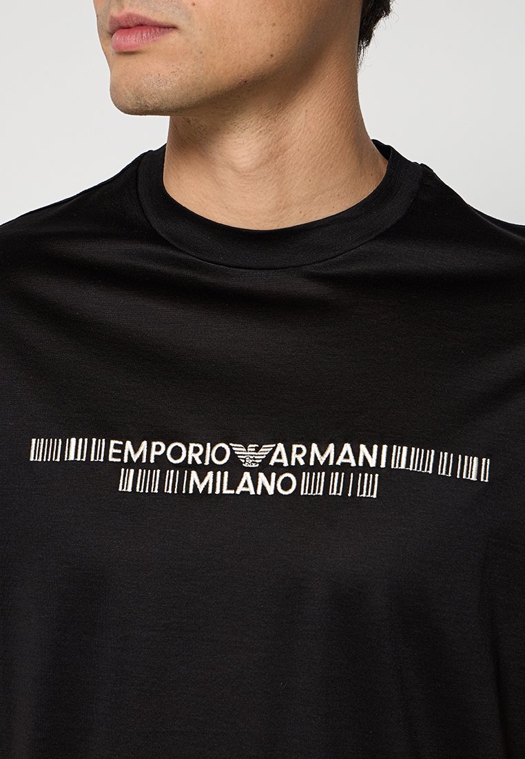 Hombre con camiseta negra con texto blanco "EMPORIO ARMANI MILANO" y logo de águila impreso en el pecho.
