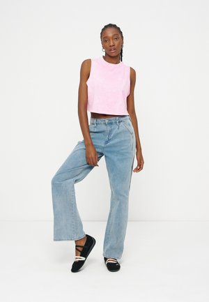 Femme portant un crop top rose sans manches, un jean large taille haute bleu clair et des chaussures slip-on noires avec des lanières blanches, debout devant un fond blanc.