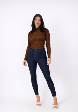 Vrouw met lang zwart haar, gekleed in een doorzichtige top met bruin patroon, donkerblauwe high-waisted skinny jeans en witte open-toe hakken, staand tegen een witte achtergrond.