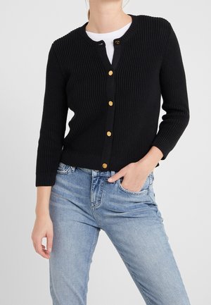 Femme portant un cardigan en maille noire avec des boutons dorés, une chemise blanche en dessous et un jean bleu clair, main dans la poche, sur un fond uni.