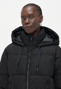 Čierna puffa bunda s vysokým golierom a nastaviteľnou kapucňou. Má horizontálne prešívanie, zips a hladkú, mierne lesklú textúru.