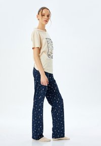 Beige Baumwoll-T-Shirt mit grafischem Druck, kombiniert mit marineblauen Schlafanzughosen mit wiederholtem himmlischen Muster und beigen Socken.