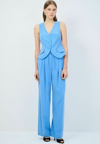 Jasnoniebieska bezrękawnik z guzikami i dwiema kieszeniami w połączeniu z szerokimi spodniami. Gładki materiał z detalem związanym w talii. Czarne sandały na obcasie.