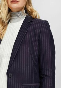 Laivastonsininen pikkutakkijakku, jossa on räätälöity istuvuus, lovetut kaulukset, kaksipainikkeinen kiinnitys ja sivutasku, päällä teksturoidun valkoisen neuleen.