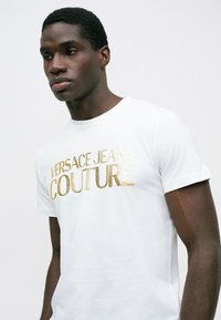 Mladý muž v bílém tričku s zlatým logem "Versace Jeans Couture", dívající se dolů a mírně vlevo, na jednoduchém pozadí.