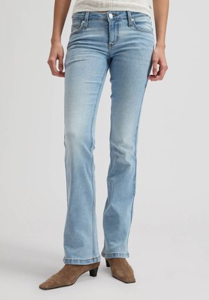 Kvinde iført lyseblå bootcut-jeans og brune ruskindstøvler, stående mod en ensfarvet grå baggrund.