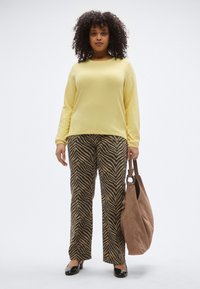 Rumena oprijeta majica z dolgimi rokavi, kombinirana z rjavimi in črnimi hlačami širokih nog. V roki drži rjavo torbico in nosi črne balerinke.