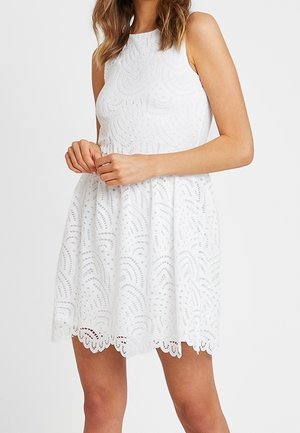 Sleeveless white lace dress with scalloped hem and intricate circular cut-out patterns, worn by a person with clasped hands.