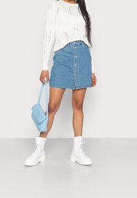 Jasnoniebieska spódnica dżinsowa z zapięciem na guziki z przodu, w zestawie z białym swetrem o fakturze dzianiny i białymi sznurowanymi botkami. Jasnoniebieska torebka na ramię.