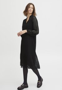 Musta laskostettu mekko, jossa on läpikuultavat pitkät hihat, V-pääntie ja pituus polveen asti. Yhdistetty tummiin loafereihin, joissa on kultaketjuaccentti.