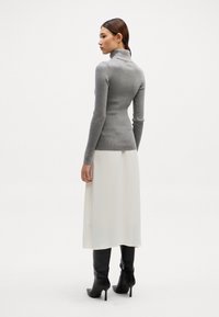 Grauer Rollkragenpullover, schmal geschnitten, gerippte Textur, kombiniert mit einem weißen Midirock und schwarzen High-Heels, von hinten betrachtet.