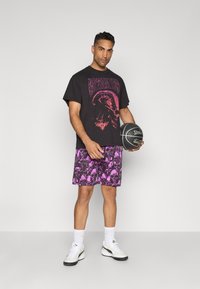 Sort grafisk t-shirt med et pink design, parret med lilla shorts med skull-mønster. Hvide sneakers og en basketball i hånden.