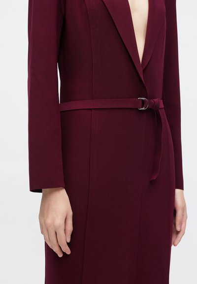 Burgundi värvi kohandatud kleit, millel on sügav V-kaelus, pikad varrukad ja sobiv vöö hõbedase pandlaga. Pehme, sile kangapind.