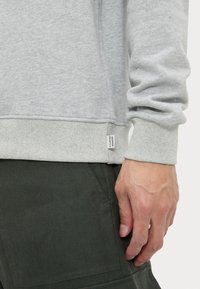 Gray sweatshirt with ribbed cuffs and hem, featuring a tag on the side. The fabric has a soft, textured finish. Hand on dark green pants.