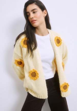 Kobieta ubrana w gruby kremowy kardigan z żółtymi wzorami słoneczników na białej koszuli i ciemnych spodniach, pozująca na jednolitym jasnym tle.