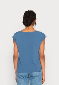 Top azul de manga corta con detalles de encaje en los hombros, que presenta un escote redondo y una tela suave. Combinado con jeans de mezclilla claros.