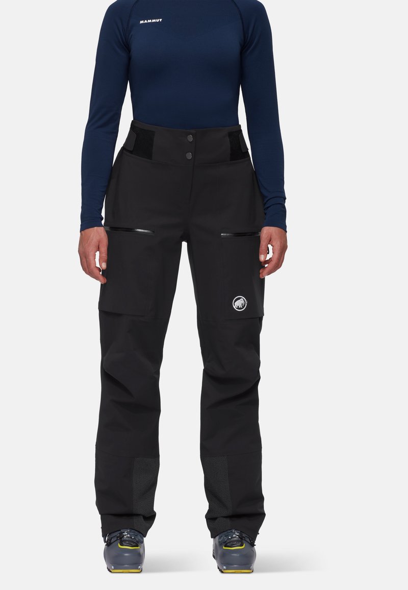 Μαύρο σακάκι σκι με φερμουάρ, πλαϊνές τσέπες, ρυθμιζόμενη μέση με βέλκρο και ενισχυμένα κάτω τμήματα. Φορεμένο πάνω από ένα σκούρο μπλε μακρυμάνικο μπλουζάκι.