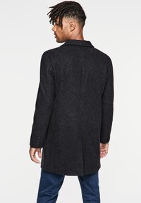 Abrigo de lana negro con corte entallado, con solapas muescas y puños de tres botones, llevado sobre un pantalón vaquero azul, visto desde atrás.