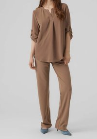 Blusa marrone leggera a maniche lunghe con colletto a incrocio e maniche arrotolate, abbinata a pantaloni su misura e tacchi azzurri chiari.