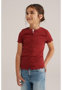 Top corto a maniche corte rosso a coste con colletto rotondo, dotato di chiusure a bottone e bordi scallop, abbinato a jeans azzurri chiari.