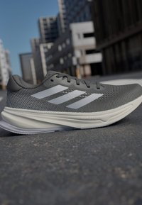 Chaussure de course pour homme avec une tige en maille grise, des rayures blanches et une semelle épaisse gris clair. Comprend des perforations pour une meilleure respirabilité.