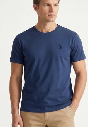 Uomo che indossa una maglietta a maniche corte blu navy tinta unita e pantaloni beige, con una mano in tasca, rivolto leggermente a destra su sfondo bianco.