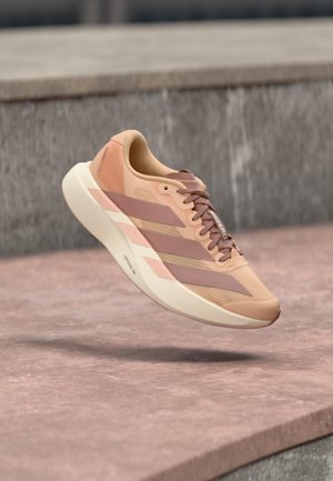 Sneaker rosa chiaro e beige con tre strisce e suola spessa, sospesa sopra una superficie di cemento rosa con uno sfondo grigio sfocato.