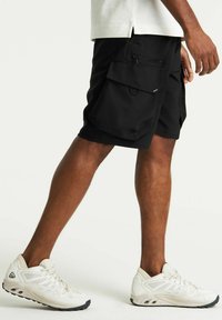 Pantaloni cargo neri realizzati in tessuto leggero, con numerose tasche, chiusura con zip e vita regolabile. Abbinati a scarpe da ginnastica bianche.