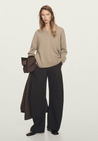 Beiger V-Ausschnitt-Pullover aus weichem Strick, kombiniert mit weit geschnittenen schwarzen Hosen. Das Model hält einen braunen Mantel. Schwarze, glänzende Schuhe runden das Outfit ab.