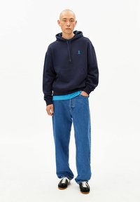 Marineblauwe hoodie met een voorzak en een blauw lintlogo, gedragen over een turquoise shirt. Gecombineerd met losse blauwe jeans en zwarte sneakers.