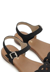 Schwarze flache Sandalen mit einem geschnallten Knöchelriemen, mit einem überkreuzten Obermaterial aus schwarzem Wildleder und einer tanfarbenen Fußbett mit Nähdetails.