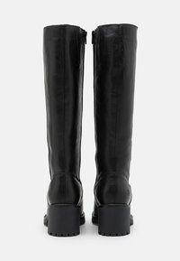 Altas botas negras de cuero con textura lisa, cremalleras laterales y tacones gruesos de goma. Las suelas presentan un patrón de dibujo para mayor agarre.