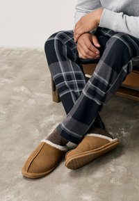Beżowe zamszowe kapcie z białym futrzanym wnętrzem, noszone z brązowymi skarpetkami i czarnymi, kraciastymi spodniami do piżamy z szarymi i jasnoniebieskimi akcentami.