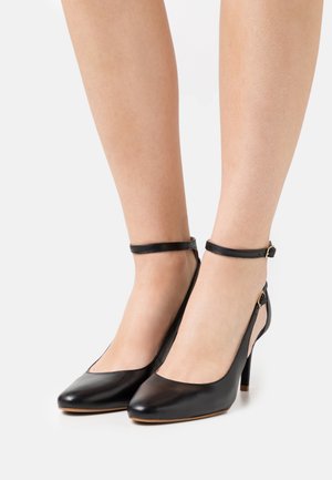 Jambes portant des chaussures à talons hauts en cuir noir avec des sangles autour de la cheville et des boucles sur un fond blanc.