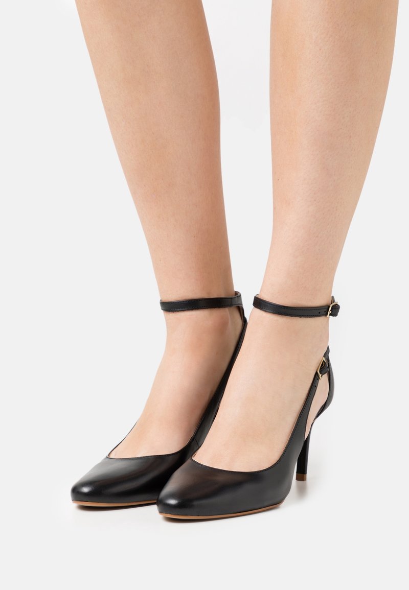 Jambes portant des chaussures à talons hauts en cuir noir avec des sangles autour de la cheville et des boucles sur un fond blanc.
