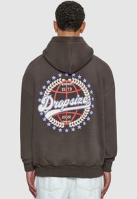 Brauner Kapuzenpullover mit Kapuze, der auf der Rückseite ein großes Grafikdesign zeigt: einen Basketball, umgeben von einem Lorbeerkranz, Text und Sternen.