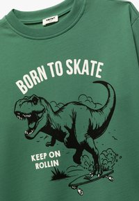 Groene sweatshirt met een ronde hals met daarop een zwart dinosaurustekeningen die aan het skateboarden is. Tekst: "GEBOREN OM TE SKATEN" en "GA DOOR MET ROLLEN."