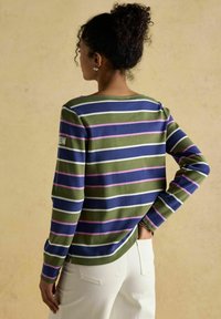 Käänud juustega naine, kellel on bun soeng, kannab pikka varrukaga roheline, sinine, roosa ja valge triibuline särk ning valgeid pükse, seistes neutraalse tausta ees.