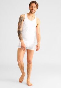 Un maieu alb din bumbac și boxeri asortați pe un model masculin. Caracteristicile includ un design ajustat și o textură netedă. Modelul stă desculț.
