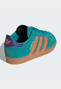 Turkos mockaskor med tre orange ränder, lila hälflik och genomskinlig gummisula. Har "Gazelle"-logotyp i guld.
