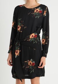 Vestido negro de manga larga, con patrones florales en rojo, naranja y verde. Detalle de cintura atada, tejido suave, longitud por encima de la rodilla.
