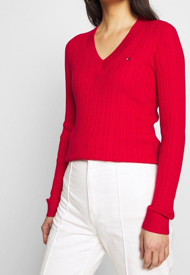 Pull-over rouge en maille avec un col en V, présentant un motif texturé. Les poignets et l'ourlet côtelés créent une silhouette ajustée. Association avec un pantalon blanc.