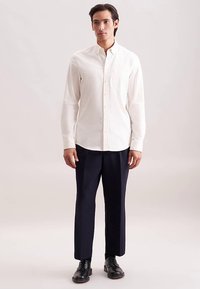 Hvid skjorte med knapper og lange ærmer, enkelt brystlomme; kombineret med marineblå bukser og sorte sko, stående mod en neutral baggrund.