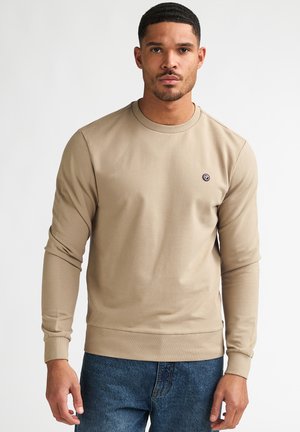 Man wearing a beige long-sleeve crew neck sweatshirt and blue jeans, standing against a plain white background.