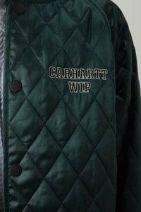 Chaqueta acolchada verde con botones oscuros y logo bordado "Carhartt WIP" en blanco y gris claro en la zona del pecho.