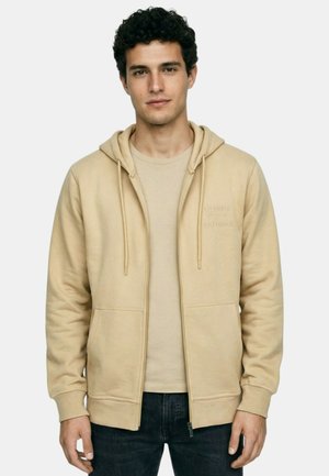 Jeune homme aux cheveux bouclés foncés portant un sweat à capuche beige zippé par-dessus un t-shirt assorti et un jean foncé, debout devant un fond uni.