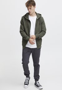 Haut de survêt votre vert avec des accents cousus, t-shirt blanc en crew underneath, pantalon sombre et baskets montantes noires, pose debout.