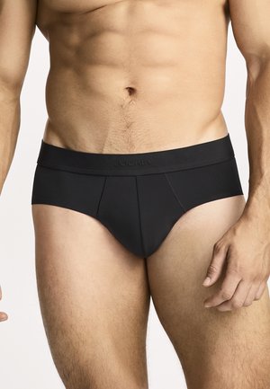 Männlicher Torso mit schwarzen Jockey-Slips, der einen definierten Bauch, Hüften und obere Oberschenkel vor einem einfarbig hellen Hintergrund zeigt.