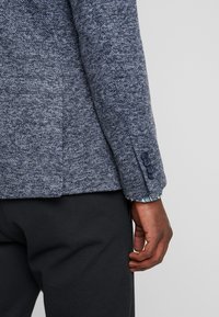 Texturerad marinblå kavajsleeve med knapppress, med en synlig mönstrad foder och skräddarsydd passform, tillsammans med svarta byxor.