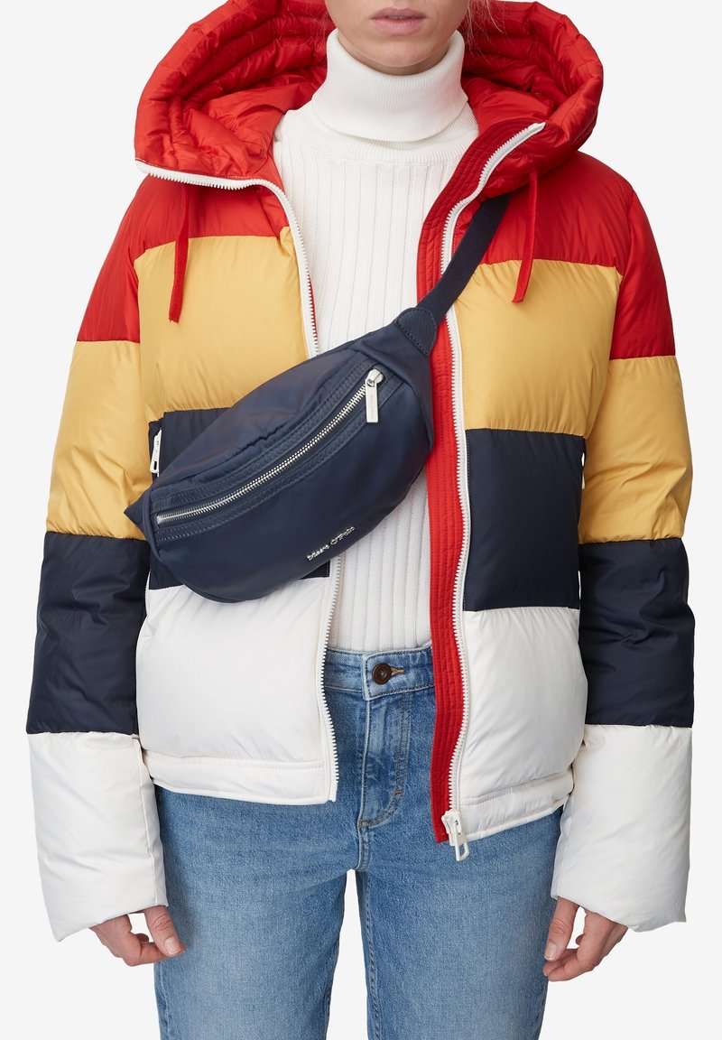 Personne portant une doudoune rouge, jaune, bleu marine et blanche, un col roulé blanc, un jean et un sac bandoulière bleu marine. Haut du corps visible.